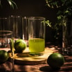 Wysokie szklanki stoją na stole. Na okrągłej desce szklanka z zielonym napojem, obok limonki i liście na ciemnym tle.