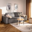 Szara sofa ustawiona na jasnym dywanie obok okrągłego stolika kawowego, z dekoracyjnym wazonem i obrazami na ścianie.