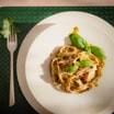 Widok z góry na talerz z szerokim, tłoczonym rantem. Na środku makaron z pesto, bazylią i kawałkami orzechów.