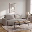 Jasna sofa ustawiona na dywanie. Przed nią stoi stolik kawowy, obok lampa stołowa. Nad sofą zawieszono grafikę.