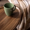 Zielony ceramiczny kubek ustawiony na owalnym stoliku w dekorze drewna. W tle widoczny jest pled.