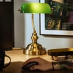 Zielona lampa biurkowa z metalową podstawą ustawiona na drewnianym blacie. W tle widoczny obraz w ramie.
