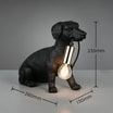 Czarna lampa stołowa w kształcie psa mającego w pyszczku kabel zakończony żarówką widoczna od przodu z wymiarami.