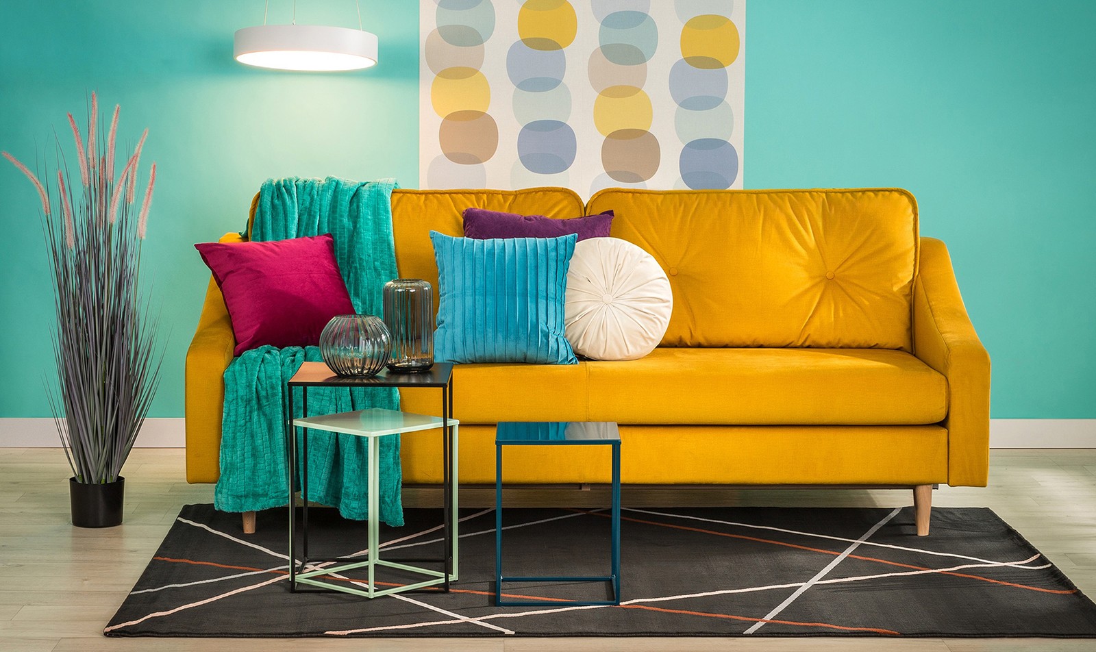 Musztardowa sofa z kolorowymi poduszkami dekoracyjnymi w nowoczesnej aranżacji salonu z turkusową ścianą.