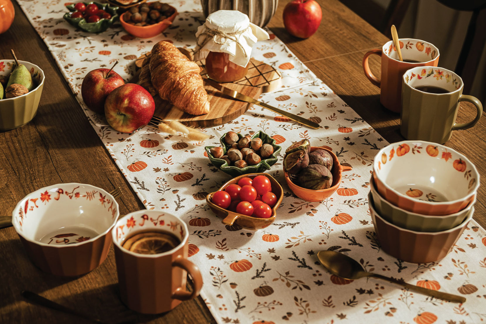 Jesiennie udekorowany drewniany stół z jasnym bieżnikiem w roślinny wzór. Na stole znajdują się ceramiczne kubki i miski, jabłka, pomidorki, orzechy, dynia oraz jesienne dekoracje w ciepłych kolorach.