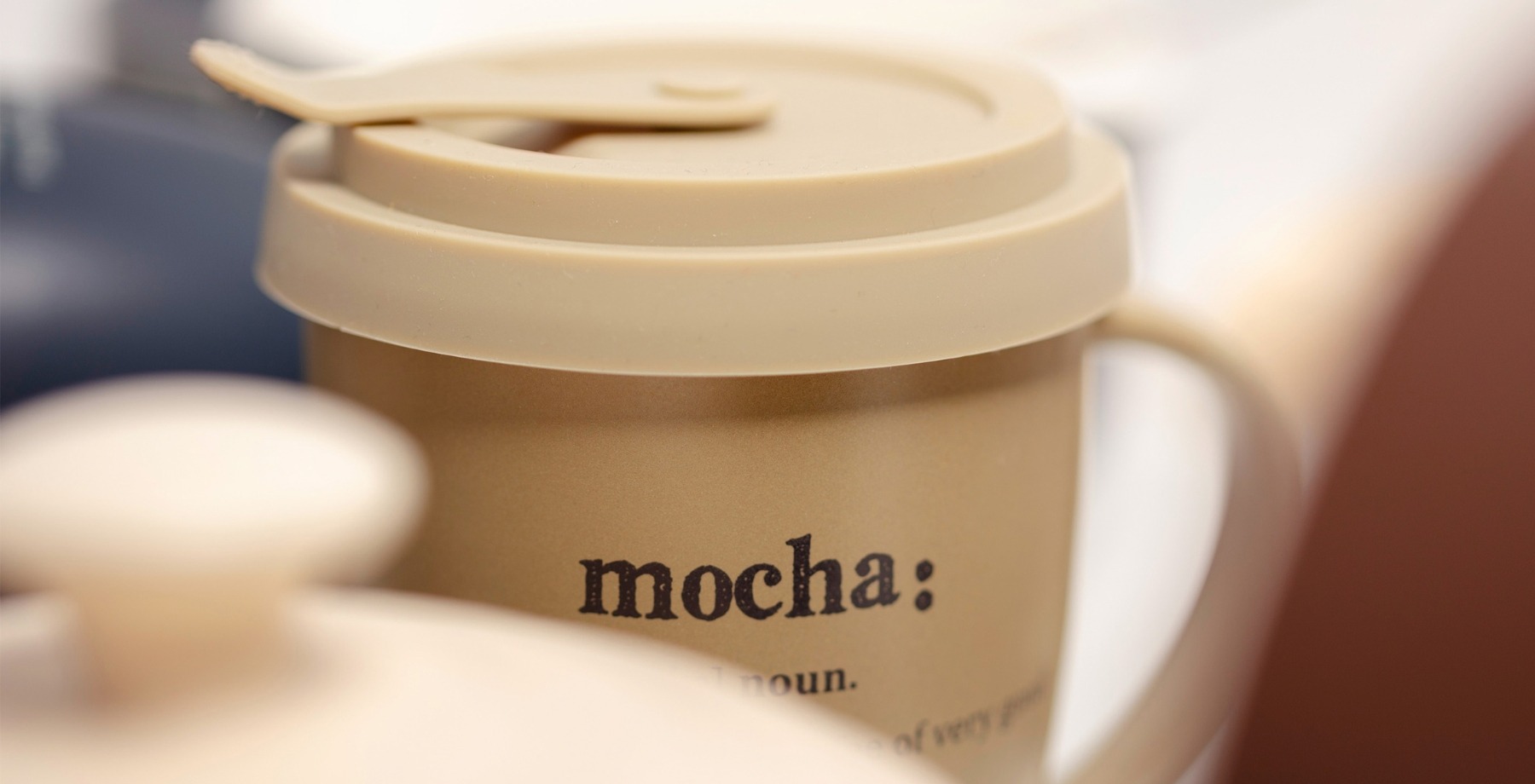 Zbliżenie na kubek z pokrywką w kolorze piaskowym. Na kubku widoczny jest dekoracyjny napis mocha