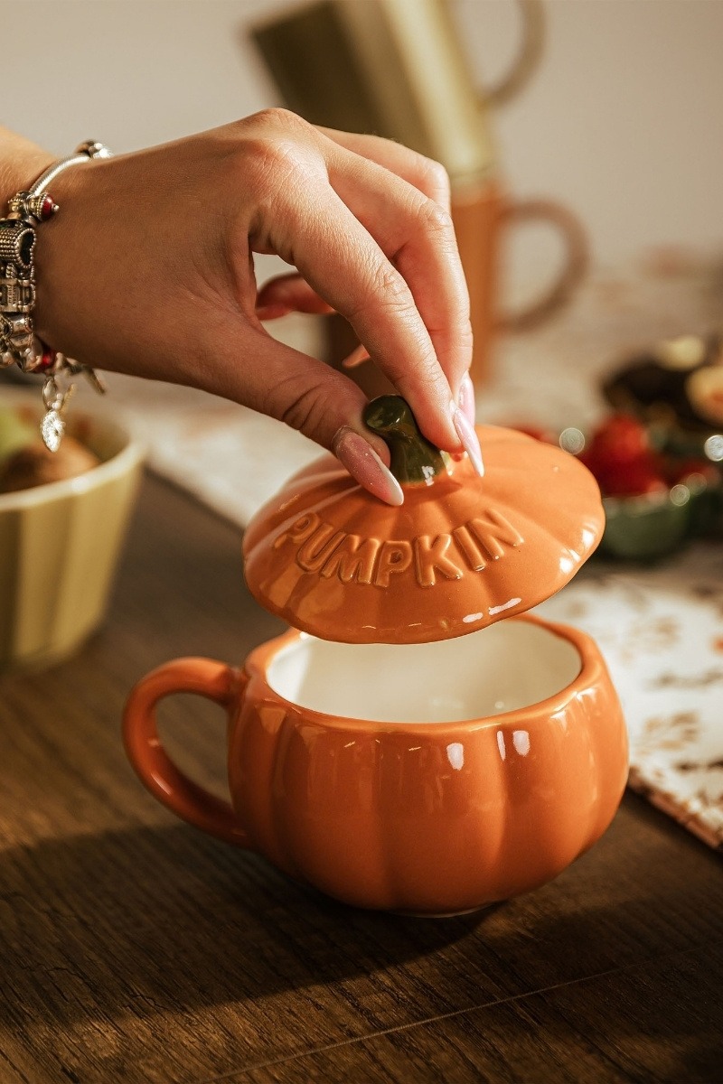 Ceramiczny kubek w kształcie dyni z pokrywką z napiszem "pumpkin" trzymaną nad naczyniem.