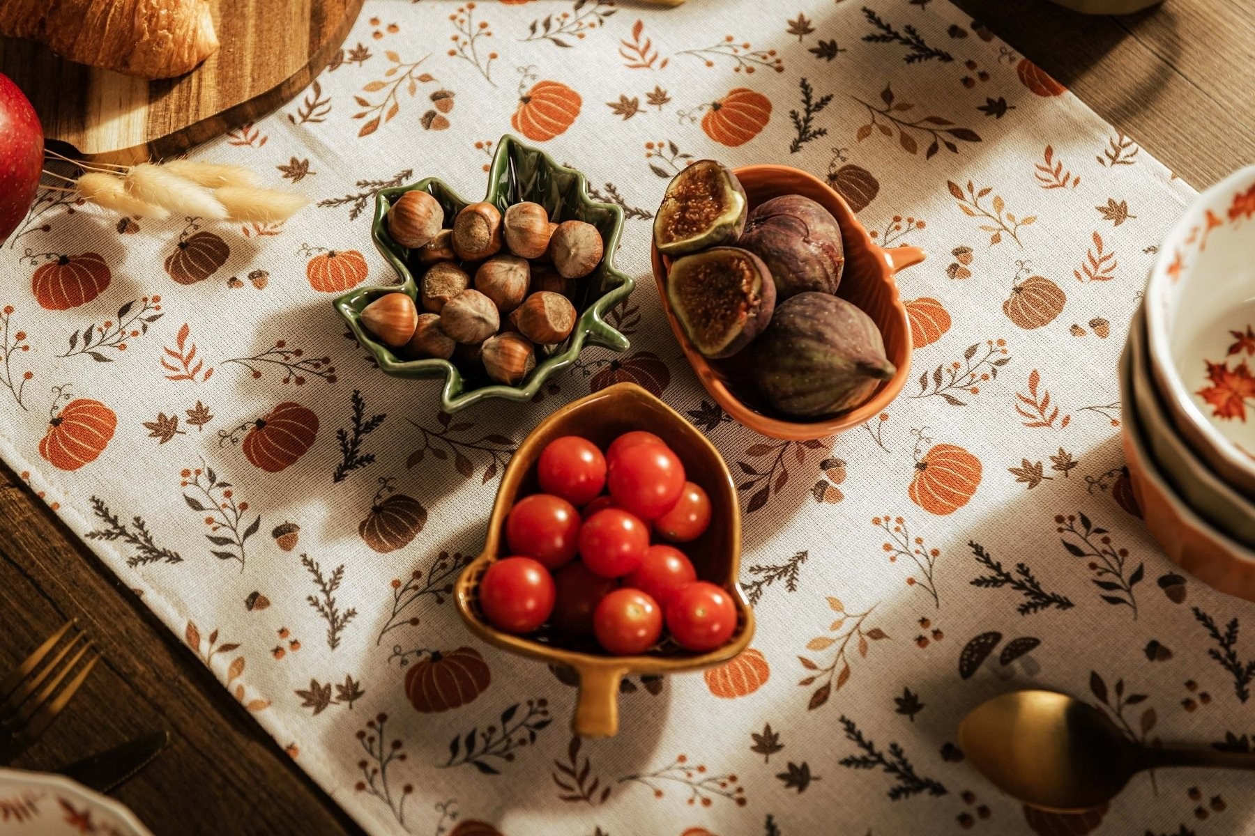 Ceramiczne miseczki w kształcie, liścia, dyni i żołędzia, wypełnione pomidorkami, orzechami i figami na jasnym obrusie z jesiennym wzorem.