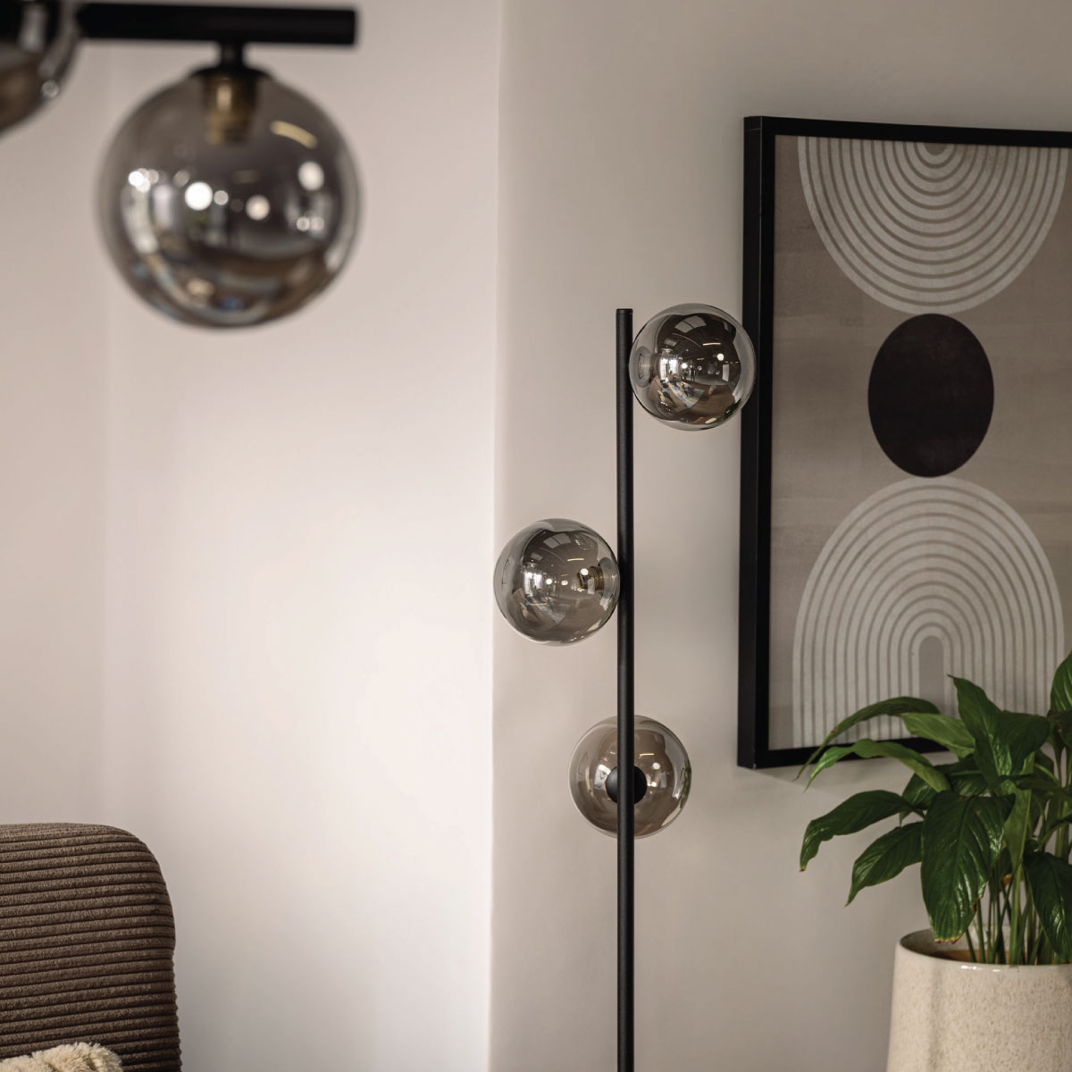 Lampa stojąca Estera z przydymionymi kulistymi kloszami obok geometrycznej grafiki ściennej i zielonej rośliny.
