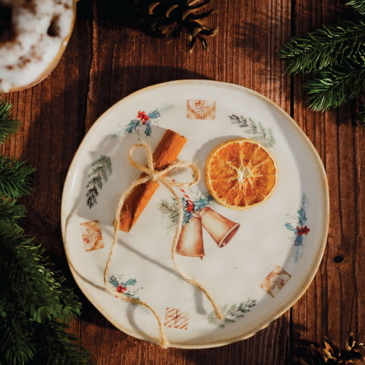 Okrągły porcelanowy talerz z delikatnym świątecznym motywem przedstawiającym pomarańcze, gałązki i dekoracyjne detale, ułożony na drewnianym stole.