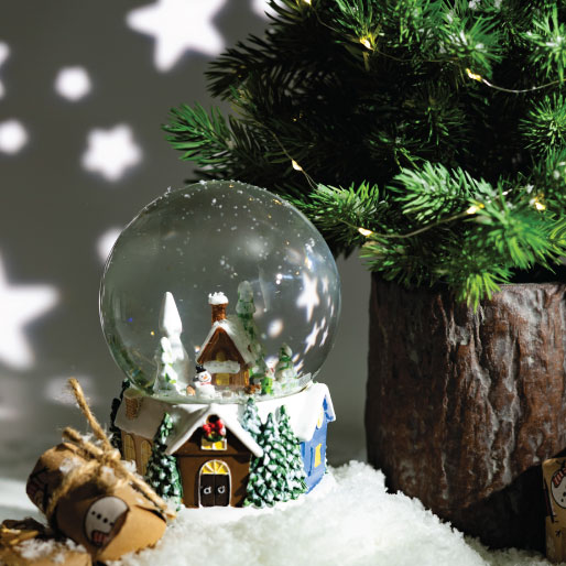 Szklana kula dekoracna z zimową scenką przedstawiającą ośnieżony domek i drzewa, ustawiona wśród gałązek iglastych i śniegu.
