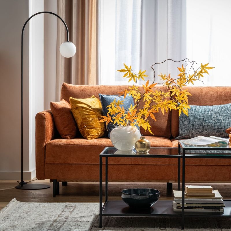 Rdzawobrązowa sofa w jesiennej aranżacji salonu, udekorowana kolorowymi poduszkami i gałązkami klonu w wazonie, ustawiona przy czarnym stoliku kawowym oraz lampie podłogowej, na tle dużych okien z jasnymi zasłonami.