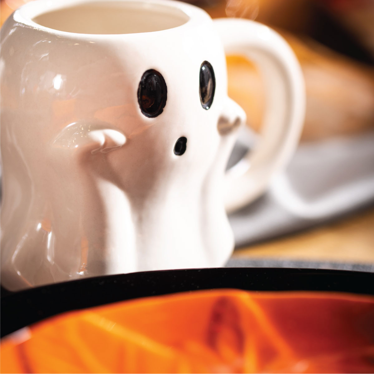 Biały kubek w kształcie ducha z czarnymi oczami i ustami, ustawiony przy pomarańczowym naczyniu
