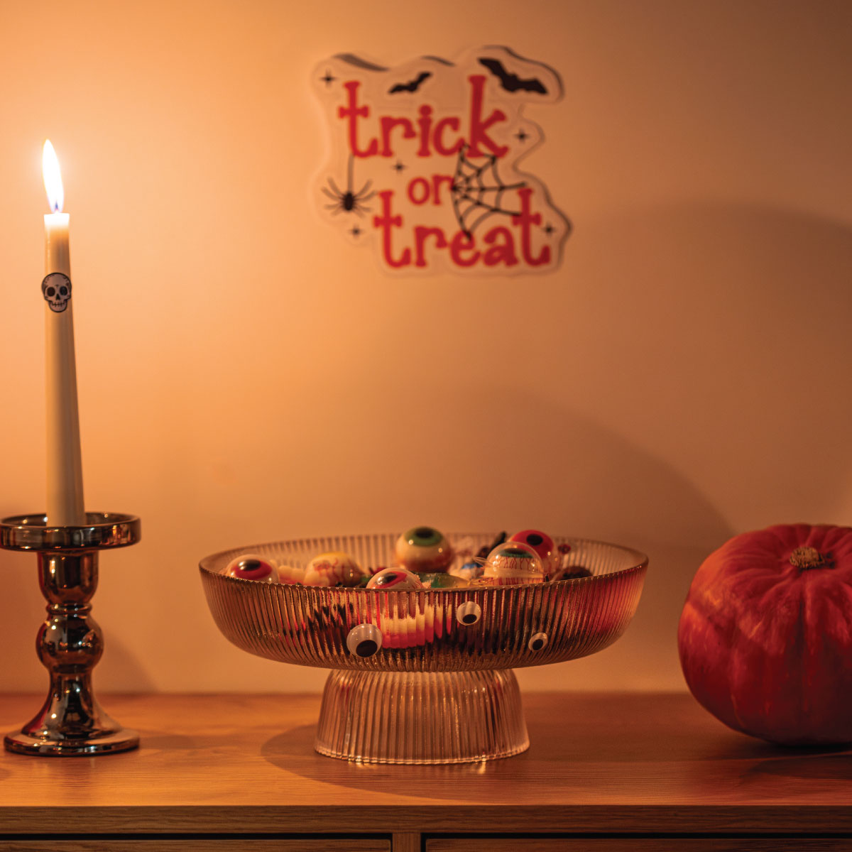 Szklana patera z przekąskami obok wysokiego świecznika i dyni, nad aranżacją dekoracja Trick or Treat