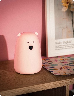 Różowa lampka TAP TAP w kształcie misia do pokoju dziecka Mała różowa lampka Tap Tap w kształcie misia stoi na regale Mauro. Obok znajduje się kolorowa książka lub zeszyt oraz oprawiona ilustracja.
