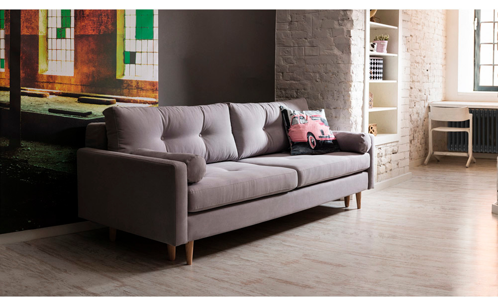 Szara sofa trzyosobowa na drewnianych nóżkach w stylu skandynawskim, ustawiona w nowoczesnym salonie z jasną ceglaną ścianą i loftowymi dodatkami.