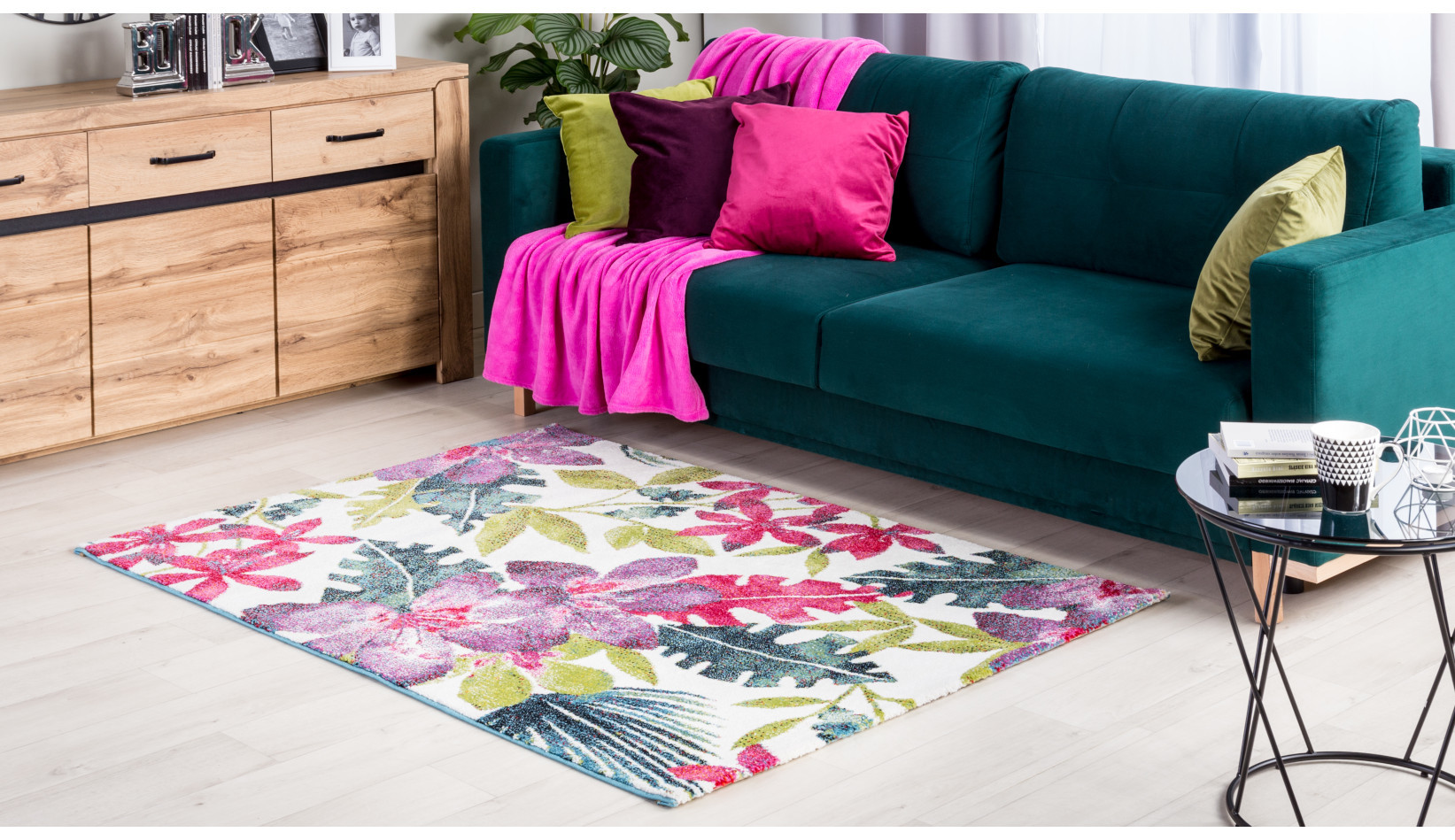 Butelkowozielona sofa z różowymi poduszkami na kwiatowym dywanie.
