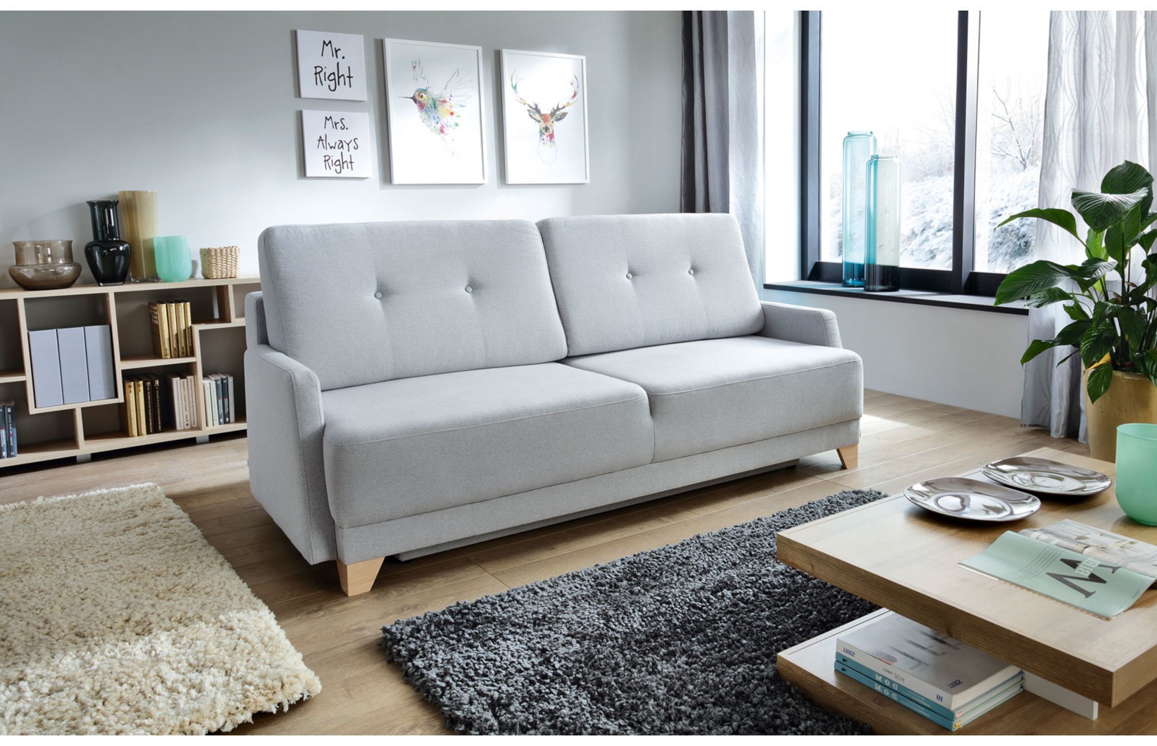 Jasnoszara sofa trzyosobowa na drewnianych nóżkach w stylu skandynawskim, ustawiona w nowoczesnym salonie z dużymi oknami i minimalistycznymi dodatkami.