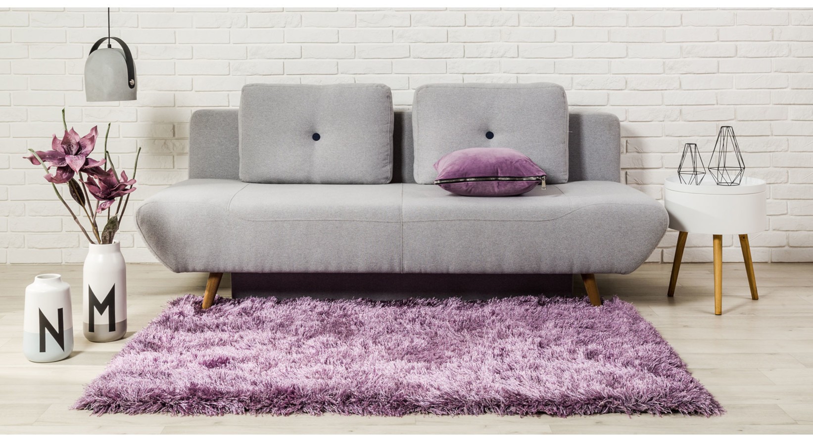 Szara sofa rozkładana na drewnianych nóżkach w stylu skandynawskim, ustawiona przy białej ceglanej ścianie z fioletowym dywanem i nowoczesnymi dodatkami.