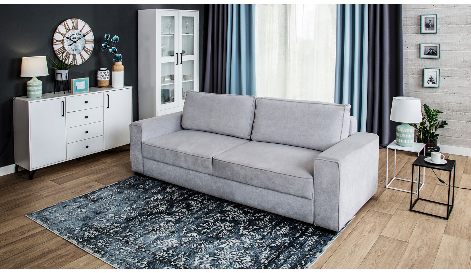 Szara sofa Elsa w nowoczesnym salonie z białymi meblami, dekoracyjnym dywanem i dużymi oknami