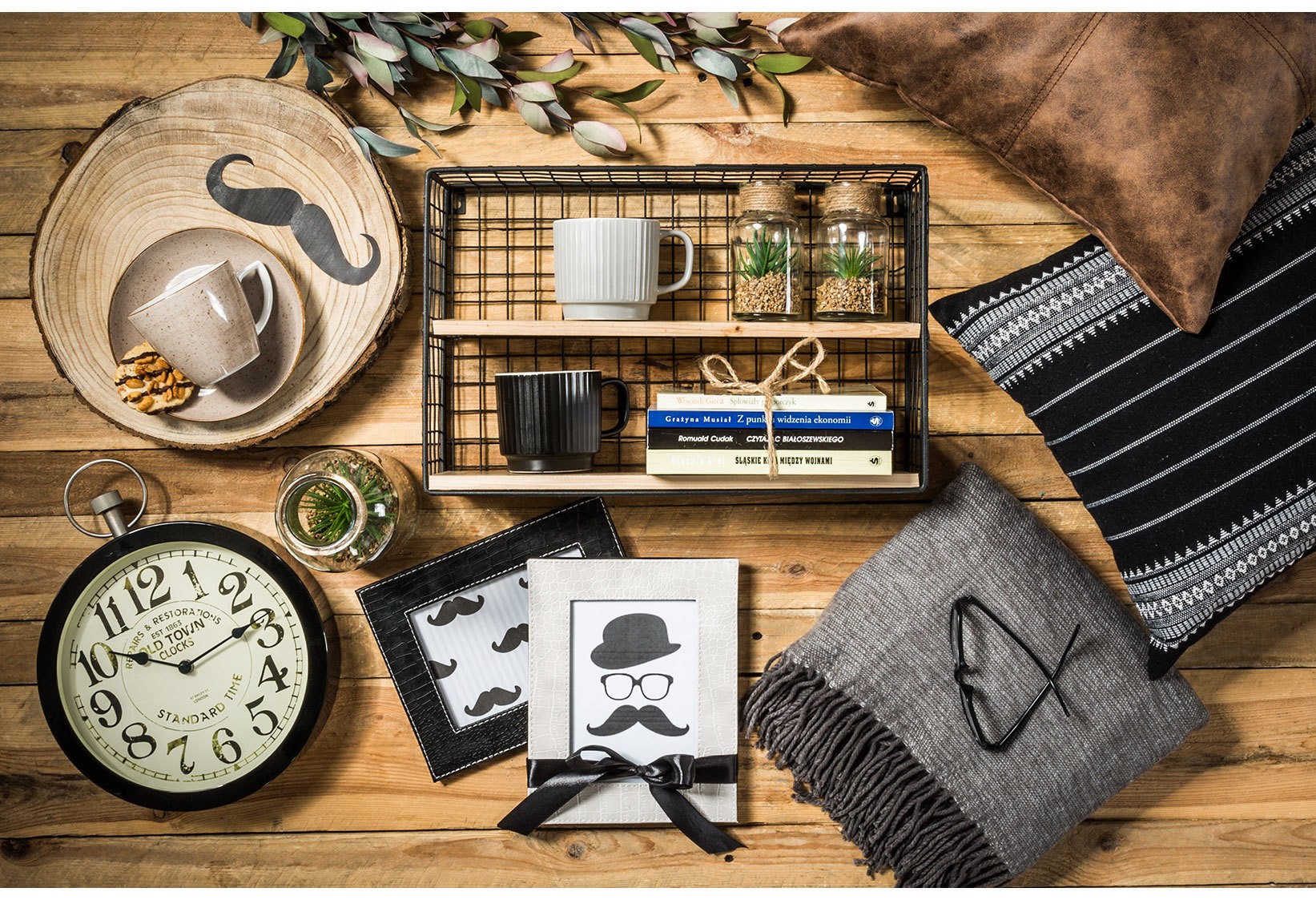 Stylowe dodatki do domu w stylu loftowym i skandynawskim, obejmujące zegar ścienny, dekoracyjne ramki, kubki, miękki pled, poduszki oraz metalowy organizer z drewnianymi półkami na tle naturalnego drewna.