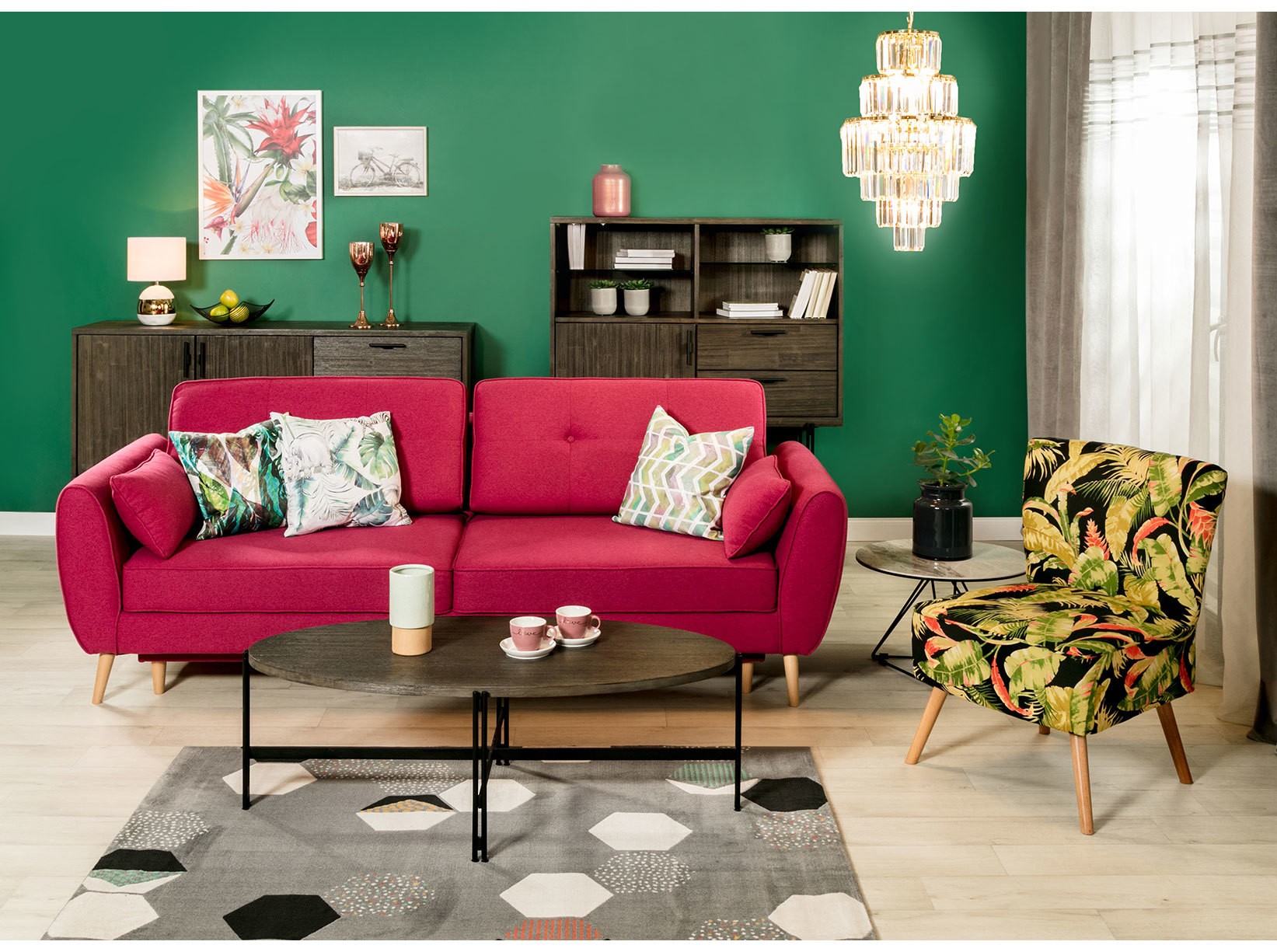 Różowa sofa trzyosobowa i fotel w tropikalny wzór w nowoczesnym salonie z zieloną ścianą, drewnianymi meblami oraz okrągłym stolikiem kawowym w stylu retro.