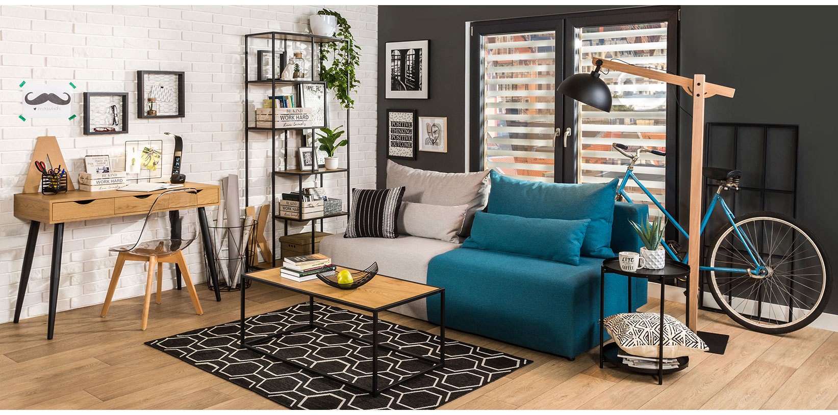 Nowoczesny salon w stylu loftowym z niebieską sofą, drewnianym biurkiem, metalowym regałem, stolikiem kawowym oraz designerską lampą podłogową, urządzony w industrialnej aranżacji z rowerem i dekoracjami ściennymi.
