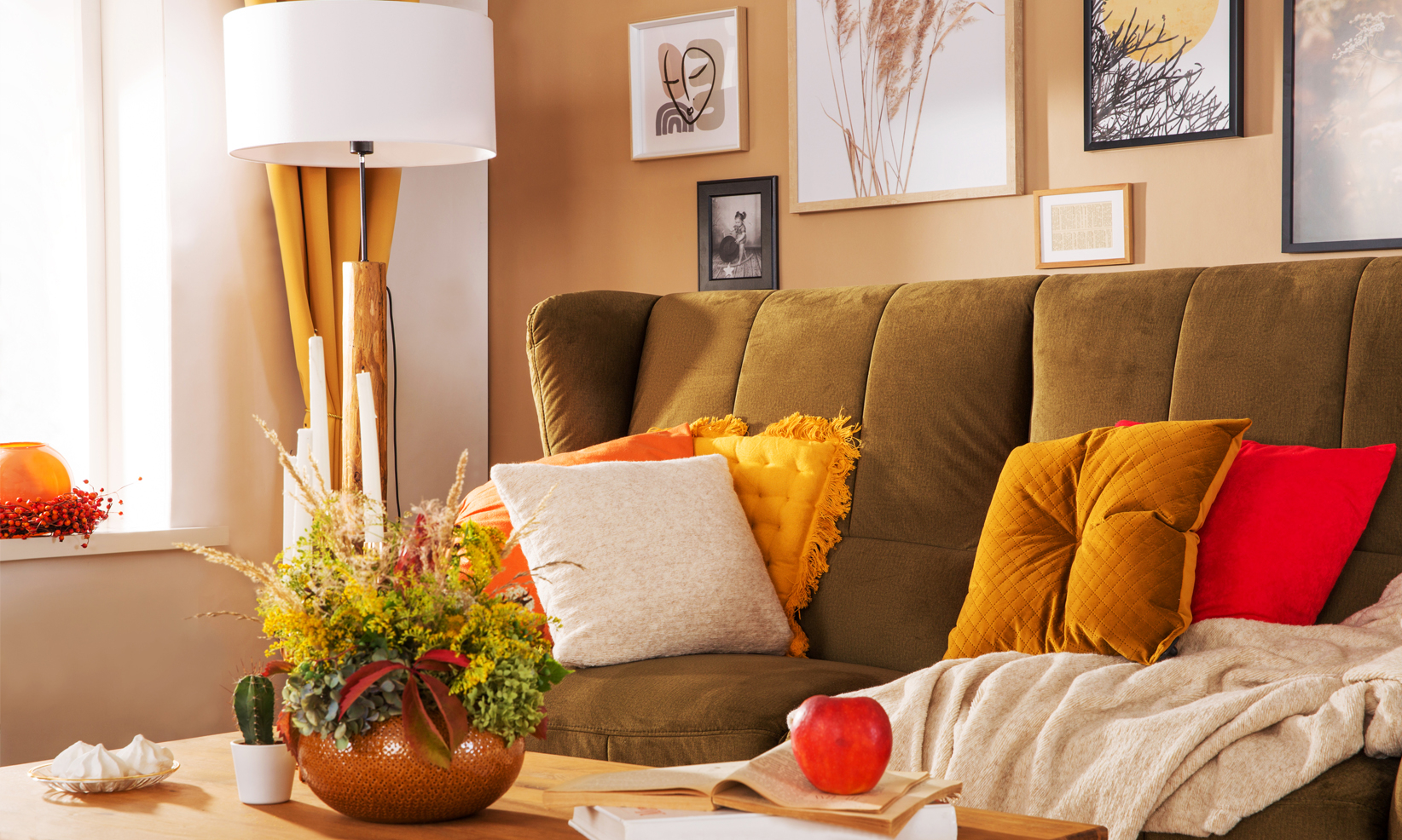 Oliwkowa sofa welurowa z dekoracyjnymi poduszkami w odcieniach żółci i czerwieni, ustawiona w przytulnym salonie z drewnianym stolikiem kawowym, lampą stojącą i jesiennymi dekoracjami.