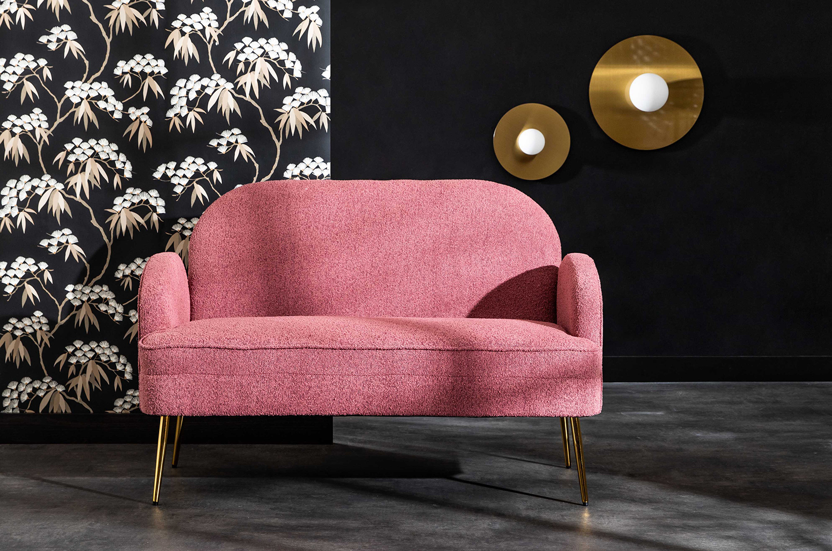 Różowa sofa dwuosobowa na złotych nogach w eleganckiej aranżacji salonu, na tle dekoracyjnej tapety w kwiatowy wzór i designerskiego oświetlenia ściennego.