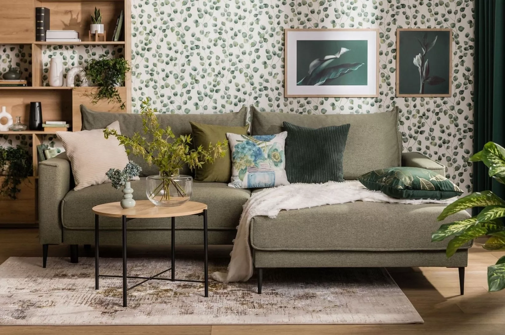 Nowoczesny salon z zieloną sofą narożną, stolikiem kawowym, drewnianym regałem oraz tapetą w roślinny wzór, urządzony w stylu inspirowanym naturą.