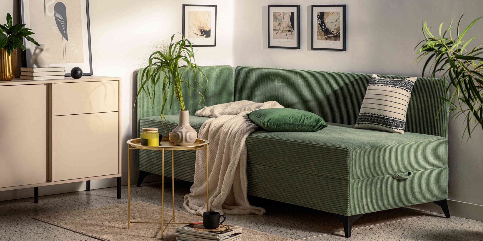 Zielona sofa z funkcją spania i pojemnikiem na pościel w nowoczesnym salonie z jasną komodą, złotym stolikiem kawowym i roślinami doniczkowymi.