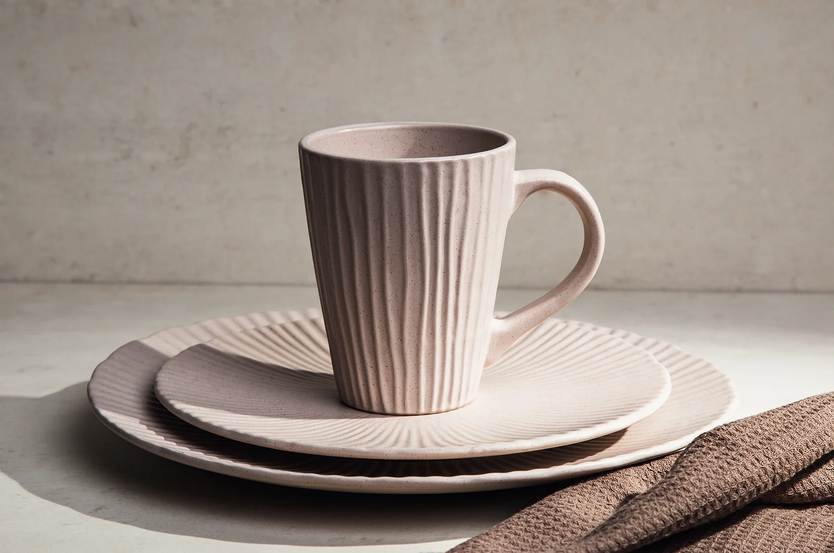 Jasny kubek ceramiczny z ryflowaną strukturą ustawiony na talerzach z tej samej kolekcji, elegancka zastawa stołowa w minimalistycznym stylu.