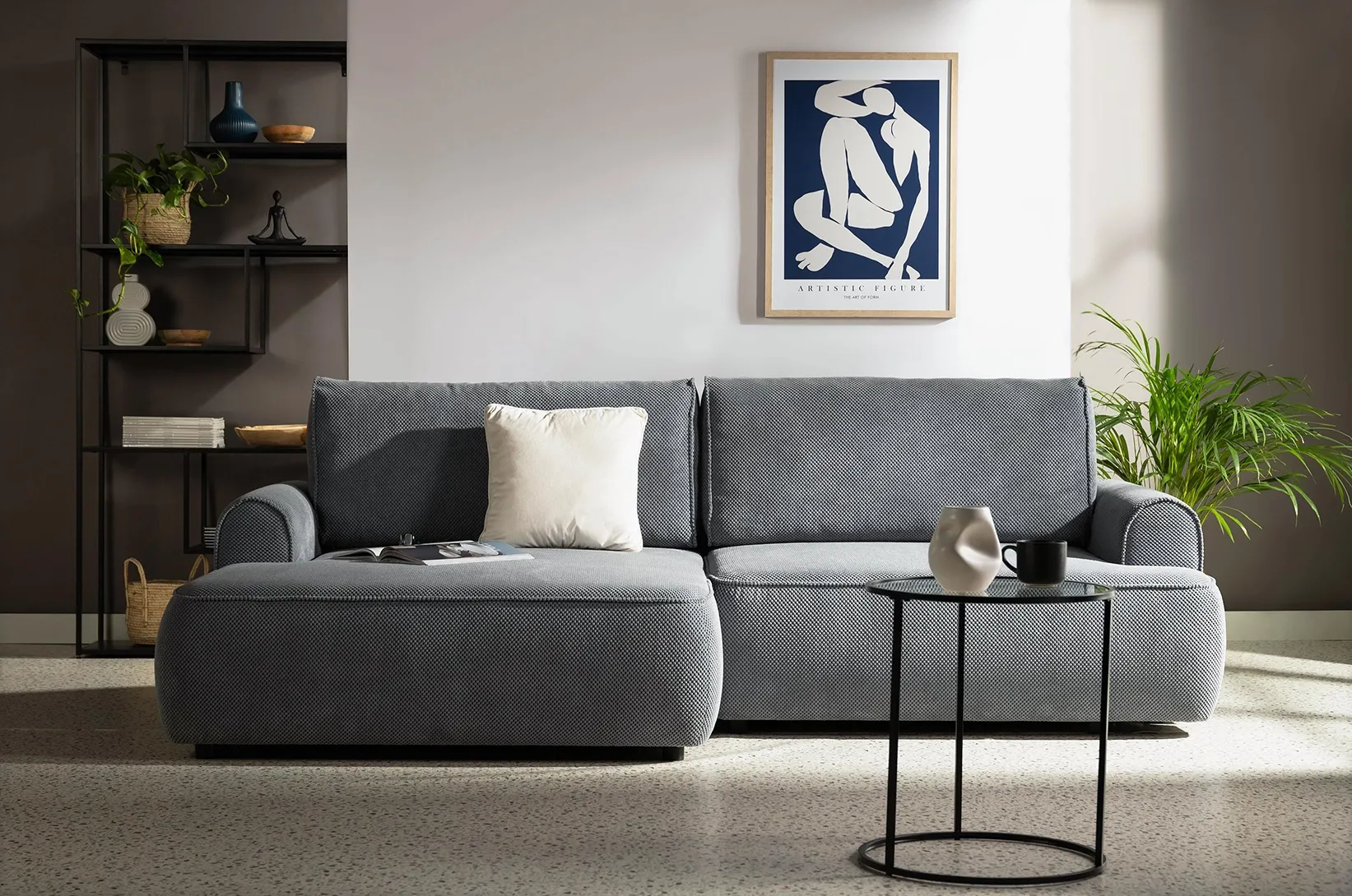 Szara sofa narożna z szezlongiem oraz czarny stolik kawowy w nowoczesnej aranżacji salonu z minimalistycznymi dekoracjami i regałem metalowym.