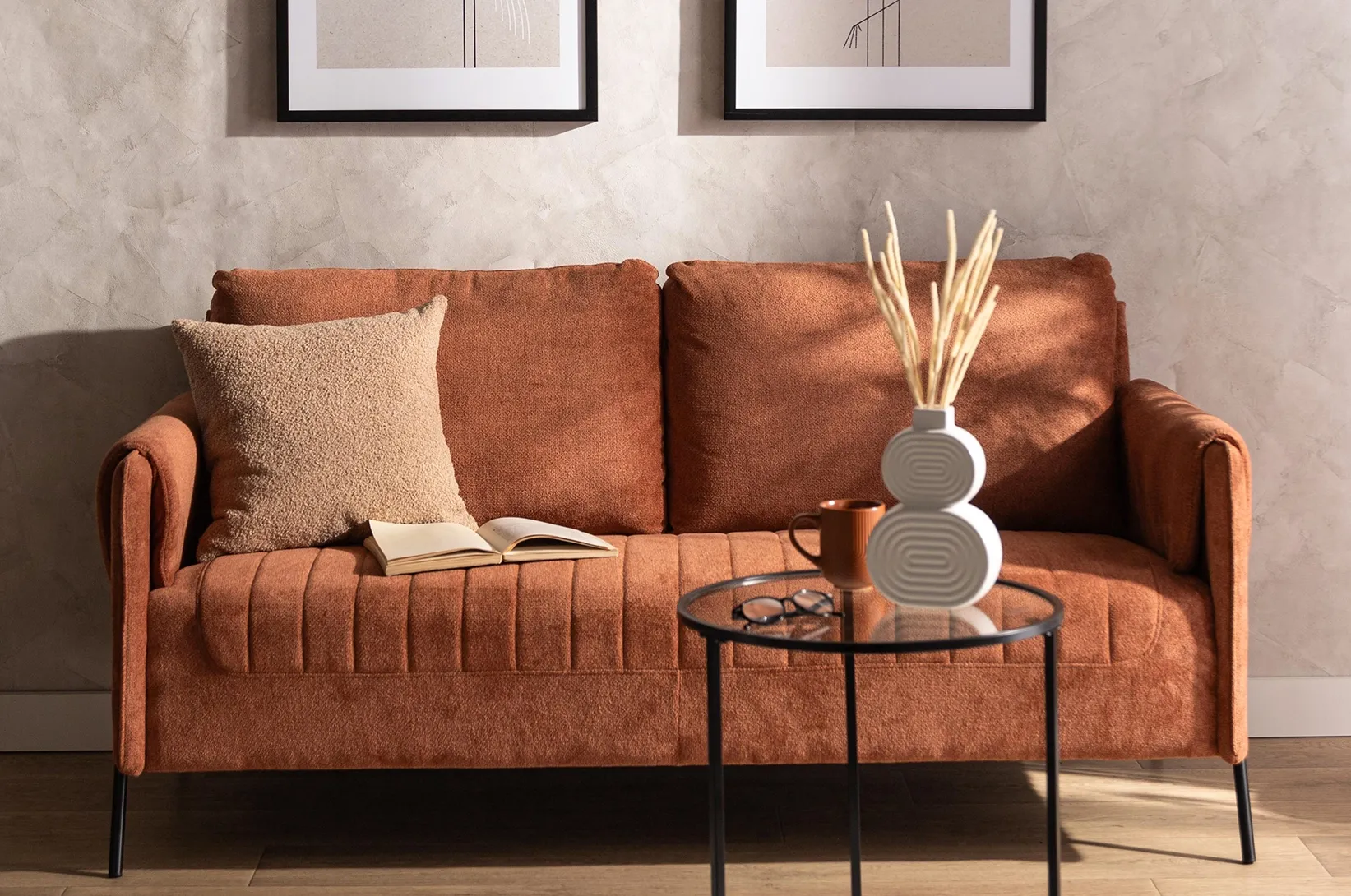 Brązowa sofa w salonie z okrągłym stolikiem kawowym, dekoracyjną poduszką i nowoczesnymi dodatkami, przytulna aranżacja wnętrza w ciepłych odcieniach terakoty.